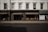 The image captures a storefront with large windows displaying various decorative items like vases and small furniture pieces. The facade has a classic architectural design with white upper walls and large, vertical windows situated above the black-framed shop displays. The street in front of the store is empty, and a billboard on the side advertises 'London Design Week'.