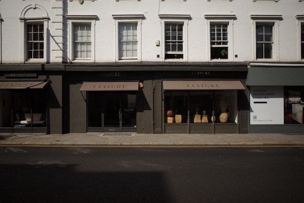 The image captures a storefront with large windows displaying various decorative items like vases and small furniture pieces. The facade has a classic architectural design with white upper walls and large, vertical windows situated above the black-framed shop displays. The street in front of the store is empty, and a billboard on the side advertises 'London Design Week'.