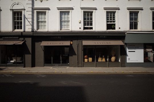 The image captures a storefront with large windows displaying various decorative items like vases and small furniture pieces. The facade has a classic architectural design with white upper walls and large, vertical windows situated above the black-framed shop displays. The street in front of the store is empty, and a billboard on the side advertises 'London Design Week'.