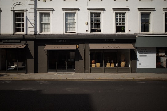 The image captures a storefront with large windows displaying various decorative items like vases and small furniture pieces. The facade has a classic architectural design with white upper walls and large, vertical windows situated above the black-framed shop displays. The street in front of the store is empty, and a billboard on the side advertises 'London Design Week'.