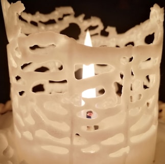 A lit candle with melted wax forming intricate, lace-like patterns around the flame. The light from the flame casts a warm glow on the surrounding wax structure.