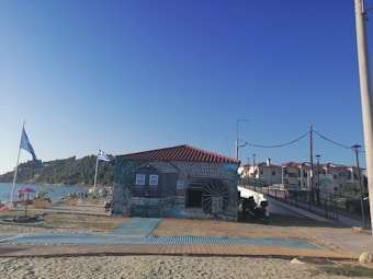 A small building with a mural depicting a watermill on its exterior wall. The structure has a red-tiled roof and is situated near a coastal area. Two flags are flying outside, one with blue and white stripes. A wooden walkway leads to the building, surrounded by sand and additional paved pathways. In the background, there are hills and a body of water, as well as several people near the shore.