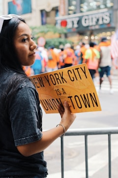 A woman holds a yellow sign that reads 'New York City is a union town' amidst a blurred background of people in vibrant clothing, suggesting a public event or gathering. A shop named 'NY Gifts' is visible in the background, indicating an urban setting.