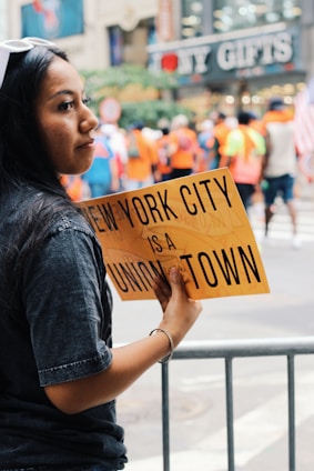 A woman holds a yellow sign that reads 'New York City is a union town' amidst a blurred background of people in vibrant clothing, suggesting a public event or gathering. A shop named 'NY Gifts' is visible in the background, indicating an urban setting.