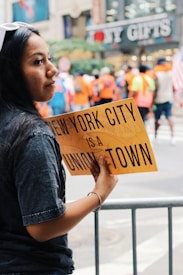 A woman holds a yellow sign that reads 'New York City is a union town' amidst a blurred background of people in vibrant clothing, suggesting a public event or gathering. A shop named 'NY Gifts' is visible in the background, indicating an urban setting.