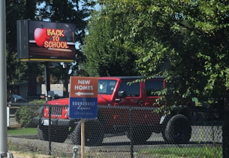 A large roadside sign displays 'Back to School' with an apple graphic. Nearby, a red SUV is parked behind a chain-link fence, partially obscured by trees. A smaller sign in the foreground advertises new homes with an arrow pointing in a direction.