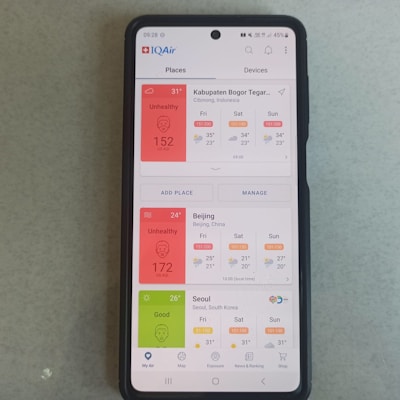 A smartphone displaying the IQAir app with air quality index information. The screen shows data for Kabupaten Bogor, Beijing, and Seoul, with varying air quality levels categorized as 'Unhealthy' for Bogor and Beijing, and 'Good' for Seoul. Weather conditions for the upcoming days are also listed.