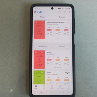 A smartphone displaying the IQAir app with air quality index information. The screen shows data for Kabupaten Bogor, Beijing, and Seoul, with varying air quality levels categorized as 'Unhealthy' for Bogor and Beijing, and 'Good' for Seoul. Weather conditions for the upcoming days are also listed.