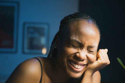 A person with braided hair is laughing with their eyes shut, head tilted slightly, and chin resting on a hand. The setting appears to be indoors, with framed pictures blurred in the background and soft lighting creating a warm ambiance.