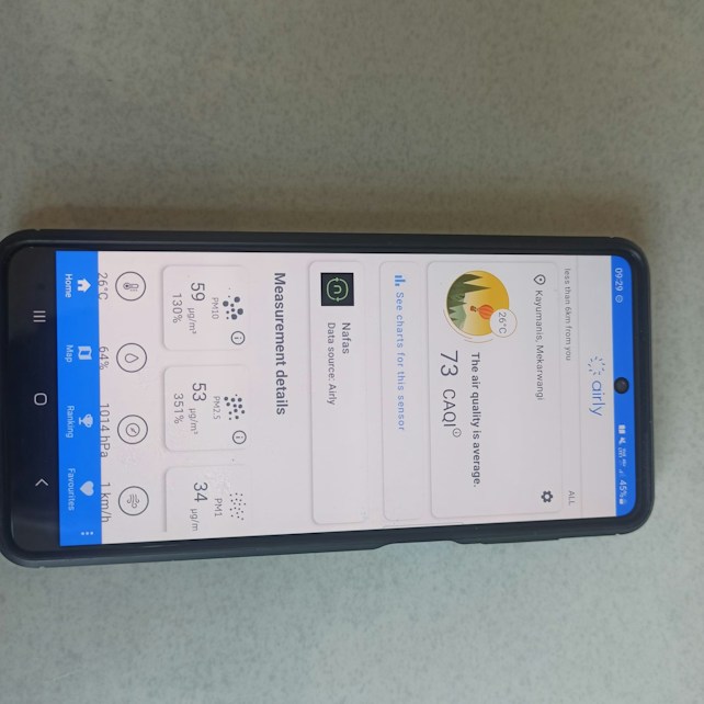 A smartphone lying on a surface with its screen displaying an air quality monitoring app. The app shows various air quality measurements in detail, including PM10, PM2.5, PM1 levels, and temperature. It also indicates that the current air quality index (CAQI) is 73 with a status of 'average'.