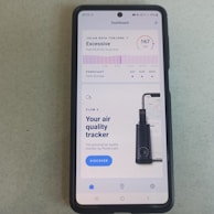 A smartphone displays an application about air quality. The screen reads an 'Excessive' air quality level with an AQI of 167. There is a chart showing a forecast for daily average air quality levels. Below, there's an advertisement for an air quality tracker device by Plume Labs with a 'Discover' button.