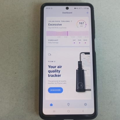A smartphone displays an application about air quality. The screen reads an 'Excessive' air quality level with an AQI of 167. There is a chart showing a forecast for daily average air quality levels. Below, there's an advertisement for an air quality tracker device by Plume Labs with a 'Discover' button.