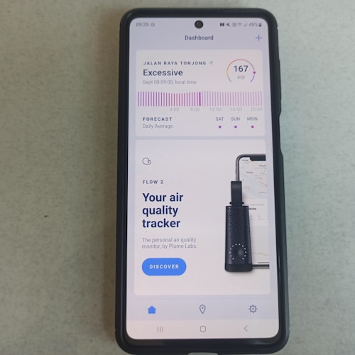 A smartphone displays an application about air quality. The screen reads an 'Excessive' air quality level with an AQI of 167. There is a chart showing a forecast for daily average air quality levels. Below, there's an advertisement for an air quality tracker device by Plume Labs with a 'Discover' button.