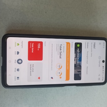A smartphone displaying an air quality monitoring app interface in Indonesian, featuring an AQI reading of 159, which is labeled as unhealthy. The screen is divided into sections with different readings and options, and the phone is placed on a light grey surface.