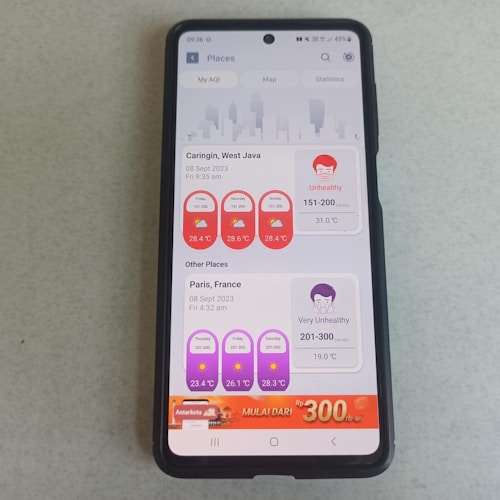 A smartphone with a black case lies on a light-colored surface. The phone screen displays an air quality application with information about different locations. The top section shows the air quality in Caringin, West Java with 'Unhealthy' status and a temperature of 31.0&deg;C. Below that, information for Paris, France shows a status of 'Very Unhealthy' with a temperature of 19.0&deg;C. There is also an advertisement banner at the bottom of the screen.