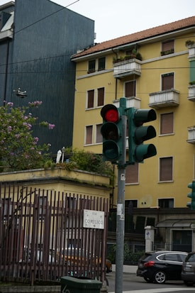 A city street corner features a traffic light showing red. A yellow residential building with balconies and green plants is visible in the background. There is a sign with 'VIA COMELICO' on a fence. Several cars are parked on the street, and a flowering tree adds a touch of nature to the scene.