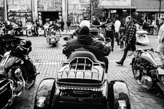 A crowded urban setting with a group of motorcyclists gathered on a cobblestone street. Several motorcycles are parked and people are milling around, some walking and others standing and conversing. Buildings with murals are visible in the background, adding a lively backdrop to the scene.