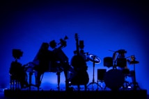 A silhouette of various musical instruments, including a drum set and a guitar, is set against a vivid blue background. The image highlights the contours and shapes of the instruments, creating an artistic and dramatic effect.