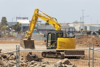 A yellow excavator is situated on a construction site with dirt piles and rocky terrain. In the background, there are industrial buildings, fences, and a partly cloudy sky. The scene conveys active construction work with machinery and equipment.