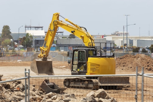 A yellow excavator is situated on a construction site with dirt piles and rocky terrain. In the background, there are industrial buildings, fences, and a partly cloudy sky. The scene conveys active construction work with machinery and equipment.