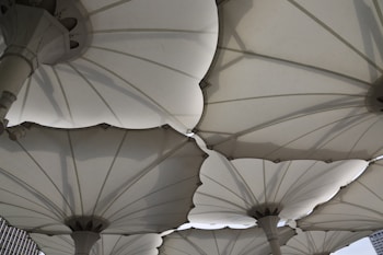 Large, white, canopy-like structures with a fabric material and mechanical support poles create a geometric pattern above. The design resembles large umbrellas or parasols with overlapping edges and visible seams. Shadows cast by the structures add depth and complexity to the scene.