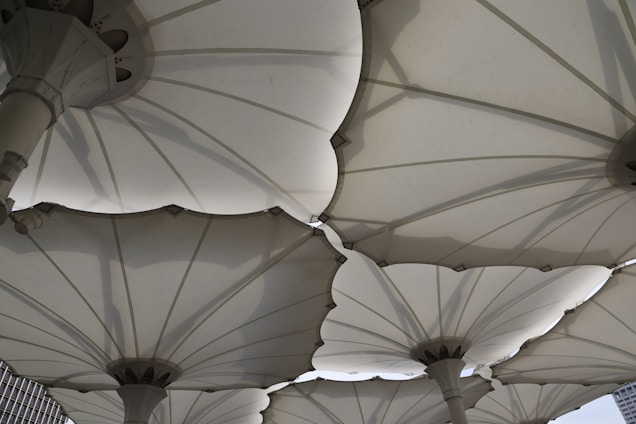 Large, white, canopy-like structures with a fabric material and mechanical support poles create a geometric pattern above. The design resembles large umbrellas or parasols with overlapping edges and visible seams. Shadows cast by the structures add depth and complexity to the scene.
