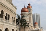 An intricately designed historical building with red domes and a clock tower, showcasing Moorish architecture. The building is accompanied by palm trees, with a modern skyscraper in the background.
