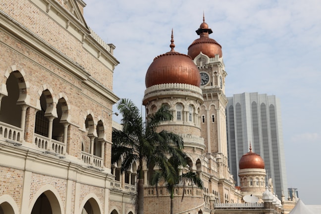 An intricately designed historical building with red domes and a clock tower, showcasing Moorish architecture. The building is accompanied by palm trees, with a modern skyscraper in the background.