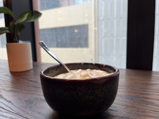 A small bowl filled with creamy yogurt is placed on a wooden table near a potted plant. The background shows large windows with faint reflections of buildings outside.