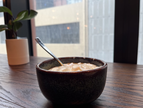 A small bowl filled with creamy yogurt is placed on a wooden table near a potted plant. The background shows large windows with faint reflections of buildings outside.