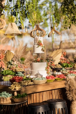 A beautifully decorated dessert table features a multi-tiered white cake adorned with flowers, surrounded by an array of colorful flowers, greenery, and decorative elements. Various hanging lamps and greenery create an enchanting atmosphere above, while wicker baskets and trays hold an assortment of treats.