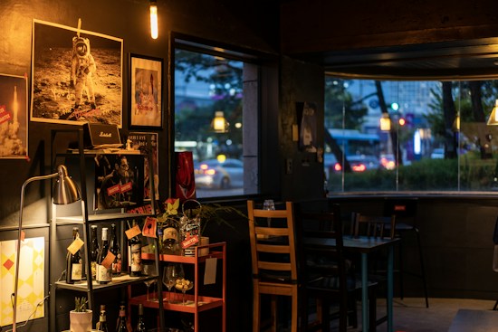 A cozy indoor scene showcasing a dimly lit room with various decorative elements. The wall displays retro-style posters, including one of an astronaut and a rocket launch. A shelf holds bottles, likely wine or liquor, and there are small plants and ornaments. A window reveals an evening city street, with blurred cars and lights visible outside.