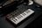 A small MIDI keyboard controller rests on a dark surface, featuring black and white keys along with various knobs and pads. The branding 'Akai MPK mini' is visible on the device. The lighting is low, giving it a professional studio ambiance.