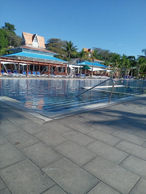 A large outdoor swimming pool is surrounded by sun loungers and tropical palm trees. The clear blue water reflects the sky and the buildings in the background, which feature unique architectural designs with triangular roofs. The area appears clean and well-maintained, ideal for relaxation.
