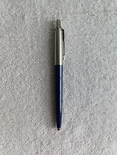 A ballpoint pen with a silver upper body and a dark blue lower body is resting on a textured gray surface.