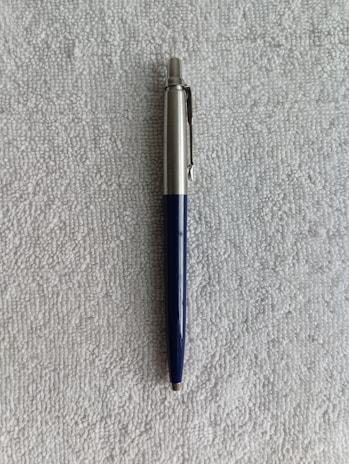 A ballpoint pen with a silver upper body and a dark blue lower body is resting on a textured gray surface.