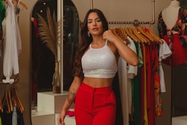 A stylish woman is dressed in a white sequined crop top and red pants, standing in a boutique or clothing store. She has long, wavy hair and is posing confidently beside a clothing rack filled with colorful garments. Nearby, there’s a mannequin wearing a floral dress, and a decorative vase with dried pampas grass adds an elegant touch to the scene.