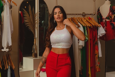 A stylish woman is dressed in a white sequined crop top and red pants, standing in a boutique or clothing store. She has long, wavy hair and is posing confidently beside a clothing rack filled with colorful garments. Nearby, there’s a mannequin wearing a floral dress, and a decorative vase with dried pampas grass adds an elegant touch to the scene.