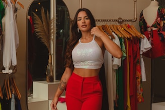 A stylish woman is dressed in a white sequined crop top and red pants, standing in a boutique or clothing store. She has long, wavy hair and is posing confidently beside a clothing rack filled with colorful garments. Nearby, there’s a mannequin wearing a floral dress, and a decorative vase with dried pampas grass adds an elegant touch to the scene.
