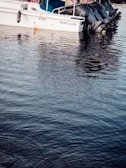 A motorboat docked on calm water, with several outboard engines. The upper part of the boat has a canopy and safety railing, while the hull displays the name 'Alhaj marine' and identification number JF-324.