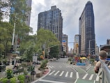 A bustling city scene featuring a prominent landmark with its unique triangular architecture. Tall buildings surround the intersection, which is lined with greenery and planters. People can be seen walking and sitting, and traffic lights guide the flow of traffic. Street signage and vibrant urban art add to the lively atmosphere.