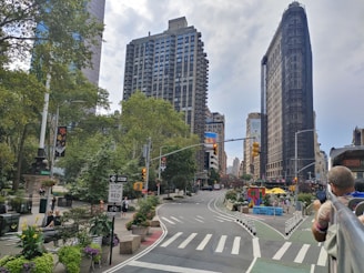 A bustling city scene featuring a prominent landmark with its unique triangular architecture. Tall buildings surround the intersection, which is lined with greenery and planters. People can be seen walking and sitting, and traffic lights guide the flow of traffic. Street signage and vibrant urban art add to the lively atmosphere.
