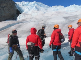 A group of people wearing bright red jackets and helmets are standing on a glacier. They are equipped with climbing gear and appear to be listening to a guide, who is gesturing and wearing a black jacket. The landscape is dominated by vast ice formations under a clear blue sky.