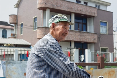 An elderly man wearing a checkered shirt and a green camouflage hat is smiling as he stands outside in front of a multi-story house with a fenced yard. The house appears modern, with large windows and a brick texture. Other buildings are visible in the background.