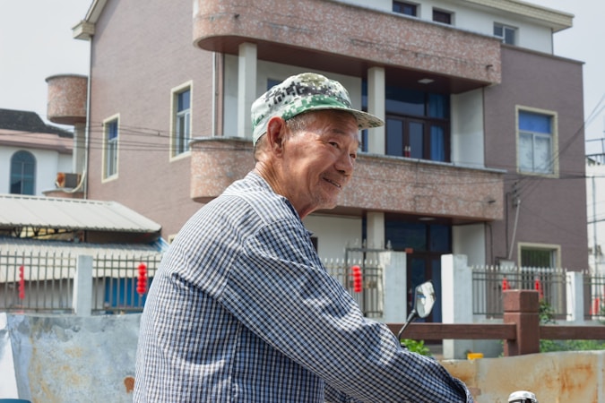 An elderly man wearing a checkered shirt and a green camouflage hat is smiling as he stands outside in front of a multi-story house with a fenced yard. The house appears modern, with large windows and a brick texture. Other buildings are visible in the background.
