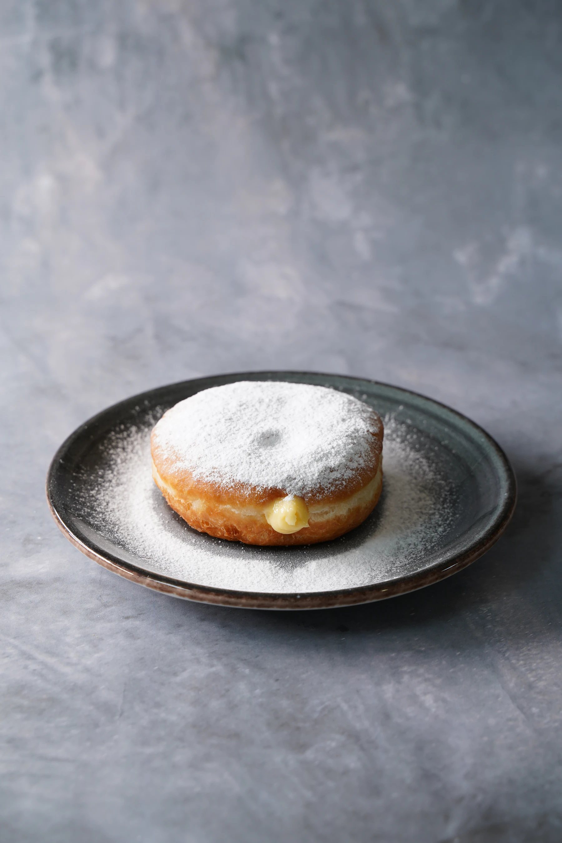 A donut dusted with powdered sugar, vanilla cream filling showing at its side, on a dark plate