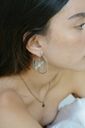 A close-up of a woman's face and shoulder wearing a large hoop earring with a butterfly design. Her dark hair partially covers her face, and she wears a delicate necklace with a small pendant.