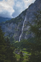A tall waterfall cascades down a steep, rocky cliff surrounded by lush green forests. Below, a small village with houses is nestled in a grassy valley. The sky is partly cloudy with dark clouds looming over the peaks of the mountains.