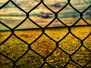 A chain-link fence in the foreground with a golden, textured water surface in the background. The sky is filled with dramatic clouds, reflecting a mix of warm and cool tones.