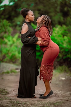 Two women stand facing each other in an outdoor setting with greenery in the background. One woman wears a black dress and has short hair, while the other wears a red dress with a lace hem and has braided hair. They appear to be engaging in a playful or affectionate interaction.